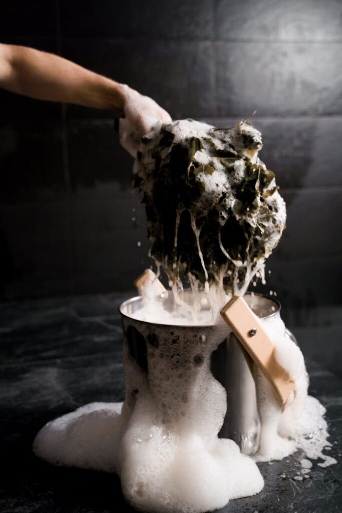A hand scrubs laundry over a bucket with soap suds and foam, highlighting cleaning and hygiene.